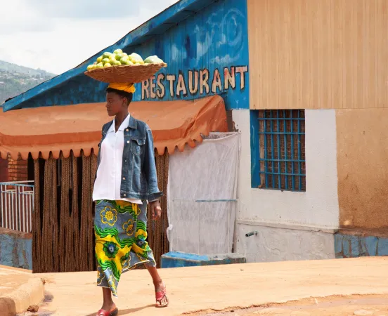 Femme portant un panier de fruits sur sa tête. Elle se tient devant un bâtiment dont une partie de la façade est peinte en bleu. Le mot "restaurant" est inscrit dessus.