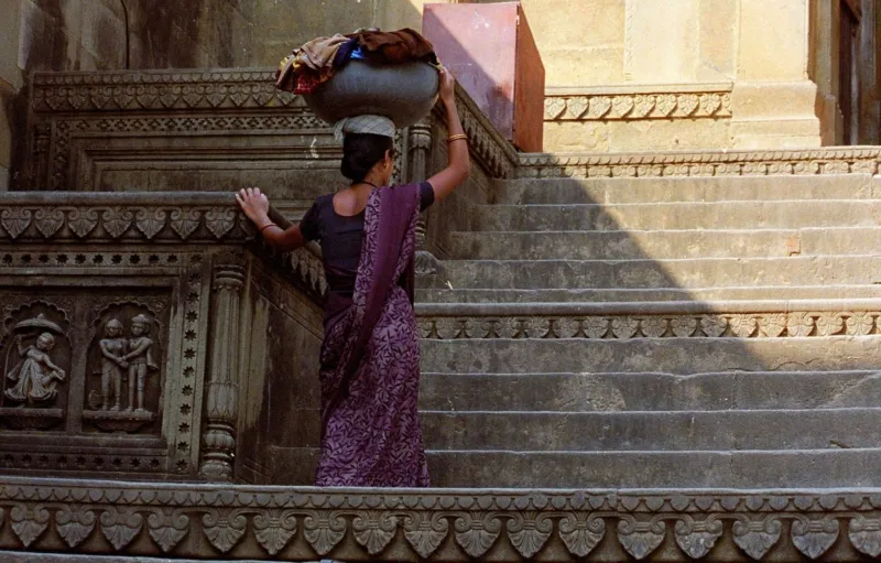 Une femme vêtue d'un sari violet porte un contenant avec des vêtements sur sa tête. Elle monte des escaliers.